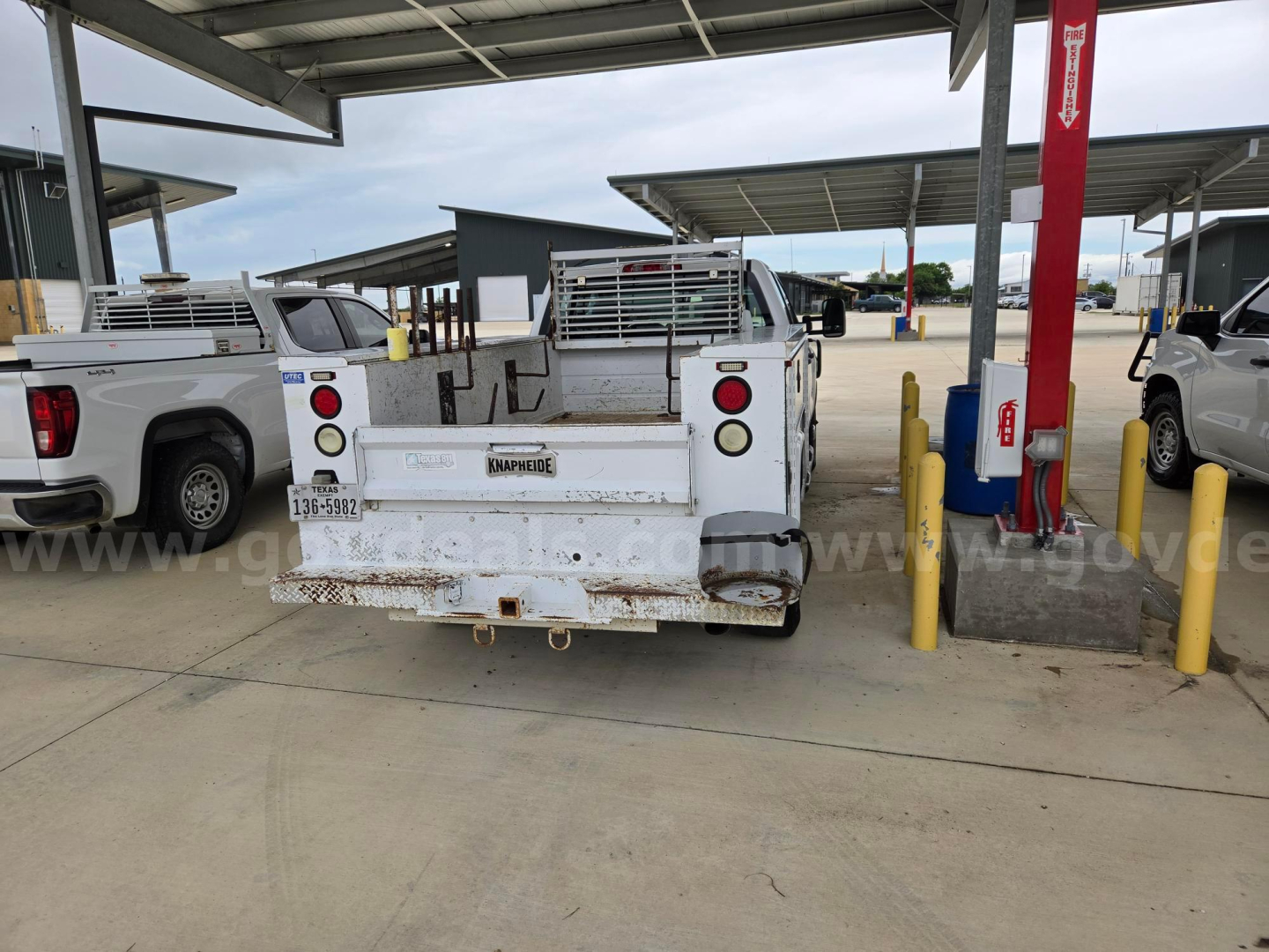 Rear view of a white work truck with a flatbed at a service center, showing tailgate with rust and trailer hitch. Visible text includes 'Transcraft' on tailgate plate, license plate area, and some badges. The surrounding area shows other tr