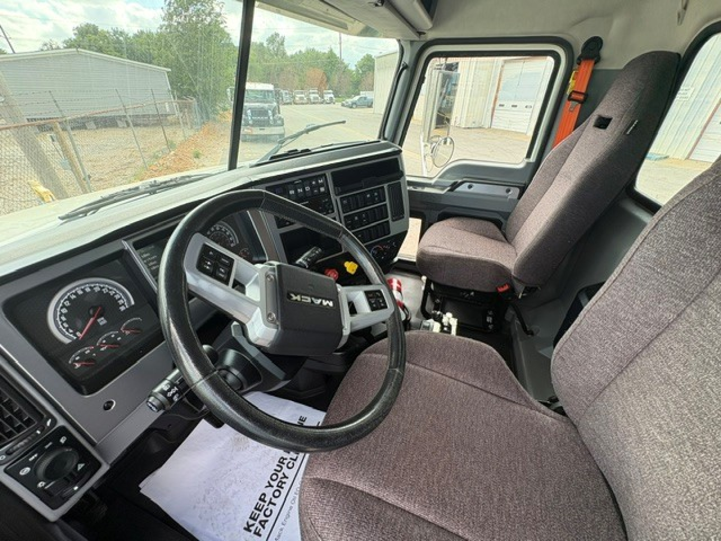 Truck cab interior with steering wheel and driver seat visible, dashboard instruments and controls, gray fabric seats, gear selector area, large windshield showing yard outside.