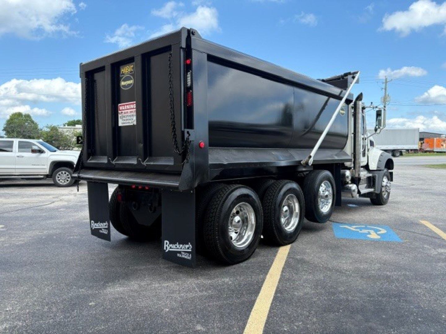Dump truck rear-right side showing six wheels, black dump bed, Mack GR84F model details not visible, warning stickers on tailgate, white cab, blue handicap parking space nearby.