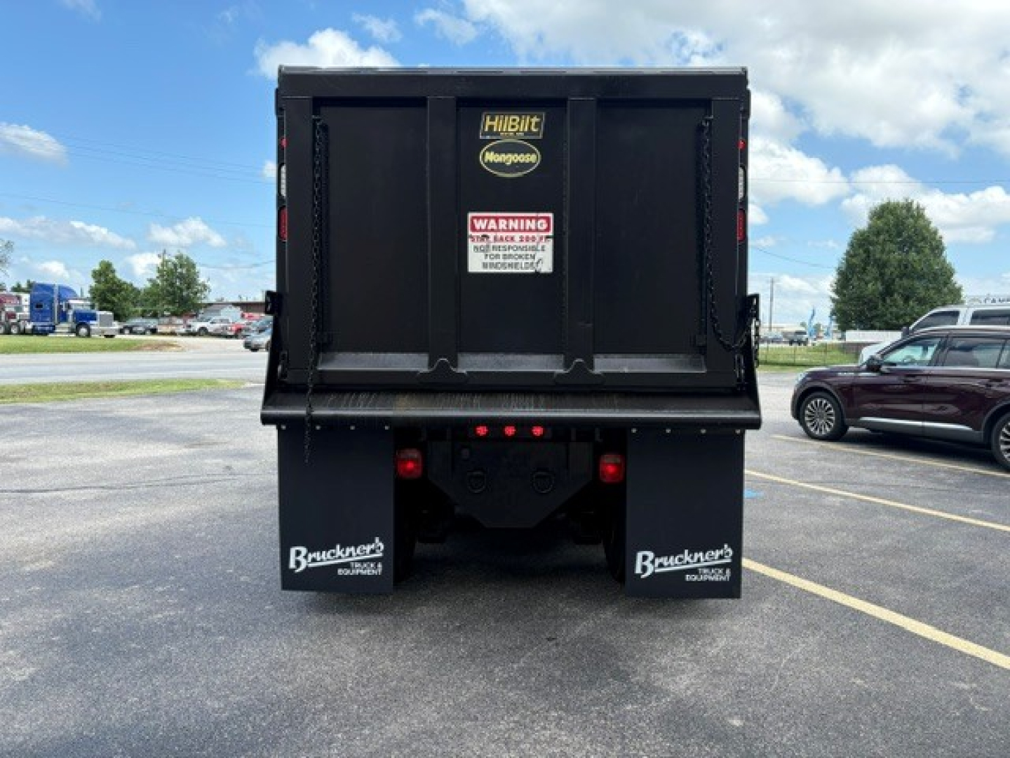 Dump truck bed rear view showing HibiIt Montgouse logo sticker, warning label, and Buckner's mud flaps. Tailgate area with black steel, truck bumper, and red brake/tail lights.