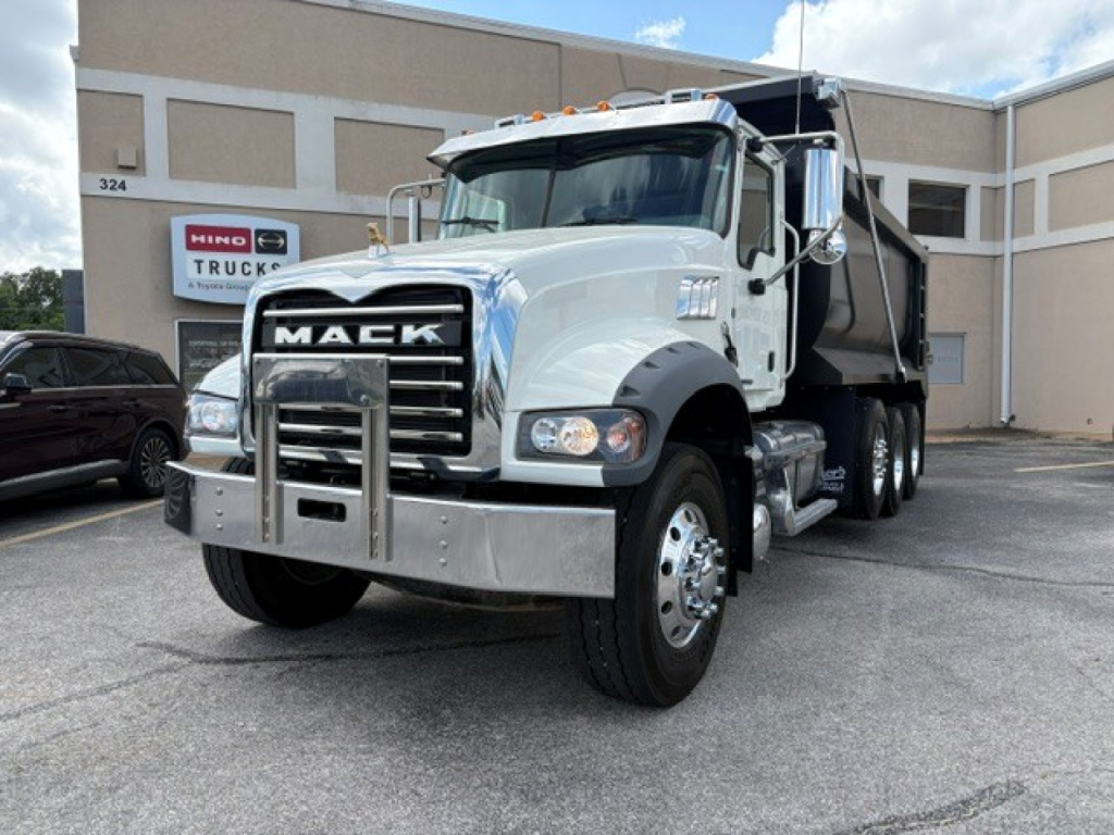 Truck front-left angle of a white 2023 Mack GR84F dump truck with chrome grille guard, dual side mirrors, and a black dump bed in a dealership lot.