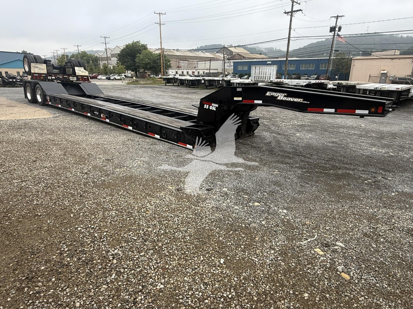 Trailer showing a long lowboy flatbed with multiple axles on a gravel lot; black with white and red reflectors, Eager Beaver branding visible on the gooseneck.