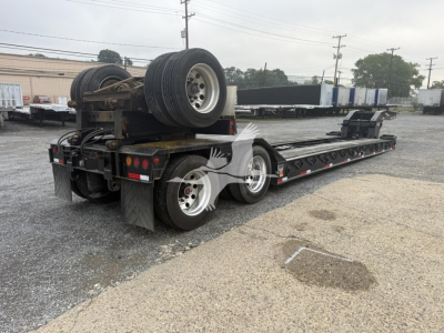 Trailer with long lowboy deck showing rear and side profile, dual rear axles, spare wheel mounted on gooseneck, and mounted fuel/air lines.