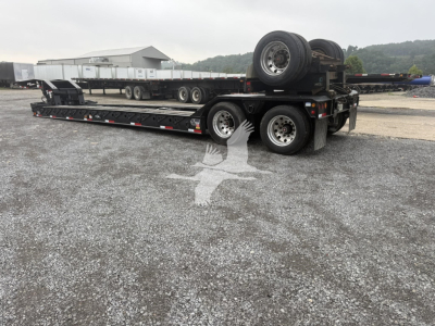Trailer showing lowboy flatbed with tandem axles, wheels, spare tire mounted at rear, and a gooseneck style prep. Visible text includes no legible labels or markings in the image.