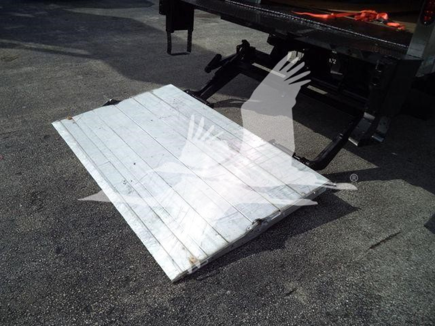 Flatbed truck with a white metal ramp lying on the ground beside a truck bed and a hydraulic lift in the background.