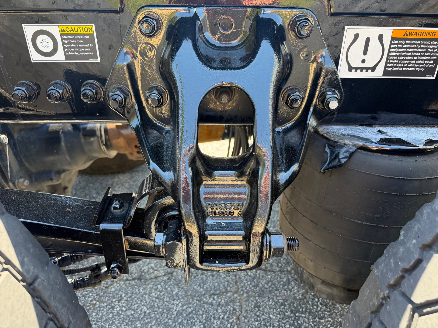 Conventional sleeper Kenworth T680 frame hitch area with bolts and warning stickers, close-up of mounting bracket and air suspension coil near wheel.