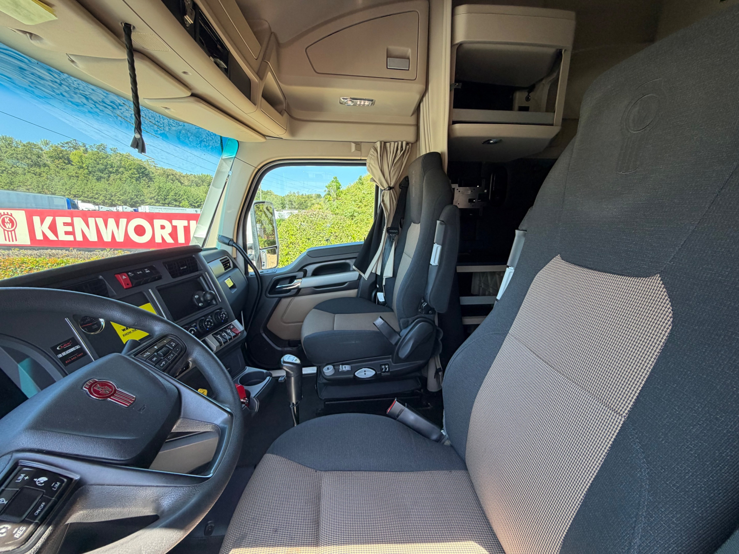 Kenworth T680 truck interior cab view showing driver seat, passenger seat, dashboard, steering wheel, and overhead sleeper area.