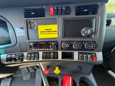 Truck dashboard with control switches, yellow warning sticker and various gauges. Visible text includes 'PLEASE WATCH YOUR SPEED', various switch labels, and a yellow key tag.