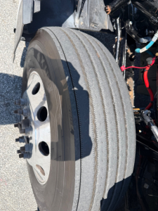 Kenworth T680 front wheel and suspension area showing tire tread and wheel hub; visible components include air lines, wiring, and frame near the engine bay.