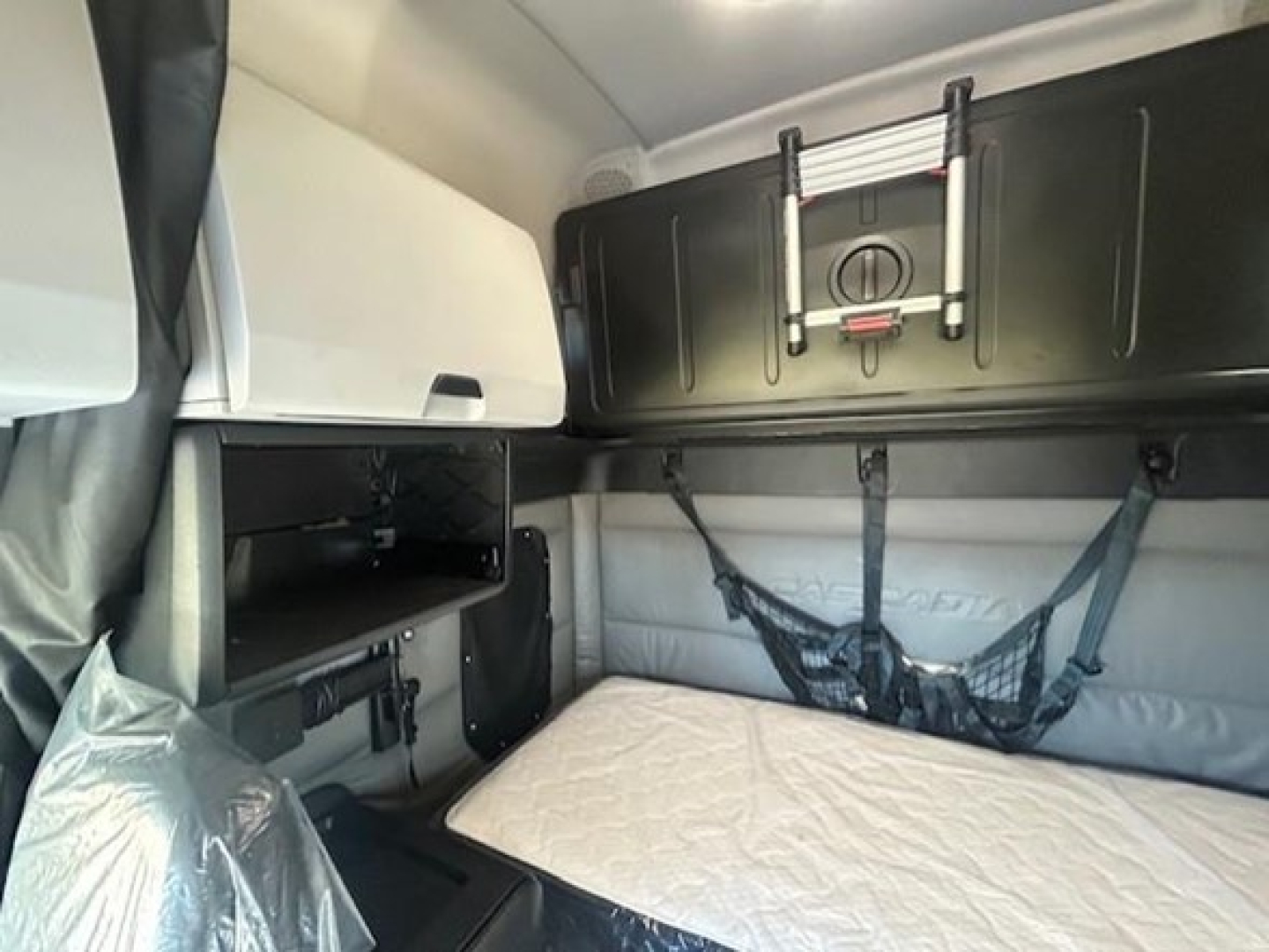 Freightliner Cascadia cab interior with sleeper bunk and storage cabinets, including a mounted overhead shelf and a bed with light-colored cover.