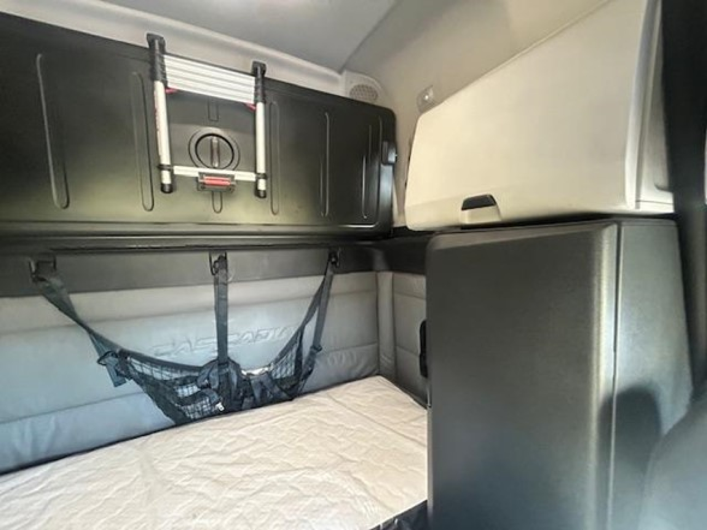 Truck cabin interior showing sleeper bed with storage cabinets and a mounted device on the upper wall of the Freightliner Cascadia cab.