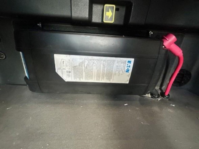 Truck cab interior showing a rectangular battery/box with white label and red positive cable connector in foreground