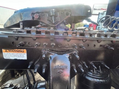 Freightliner Cascadia engine bay view showing steering area and various hoses; front of engine with hood open, branding 'Freightliner' on accessory plate; warning sticker visible.