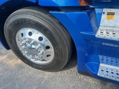 Front wheel and blue cab of a Freightliner Cascadia 126 truck showing Bridgestone tire and polished hubcaps with steps and warning label on fender.