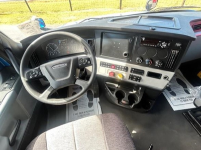 Cab interior of a Freightliner Cascadia 126 showing steering wheel, dashboard, center console with controls, and cup holders.
