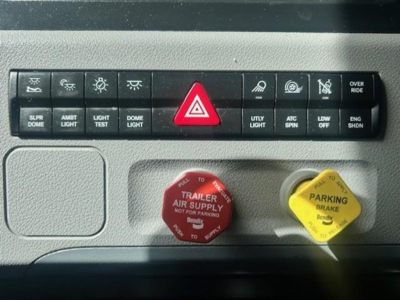 Dashboard control panel and knobs from a 2022 Freightliner Cascadia showing Hazard light button, trailer air supply, parking brake label, and various switches with small icons.