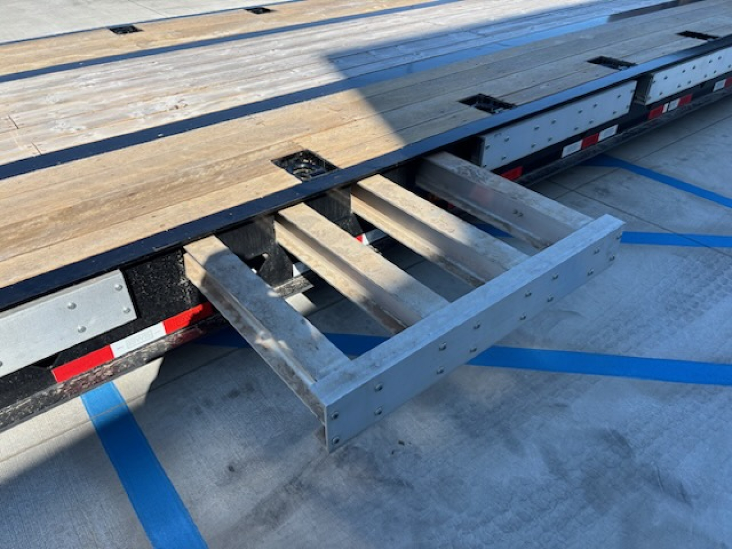 Trailer frame with wooden deck planks and metal supports; a metal ladder rung/step protrudes from the side under the deck.