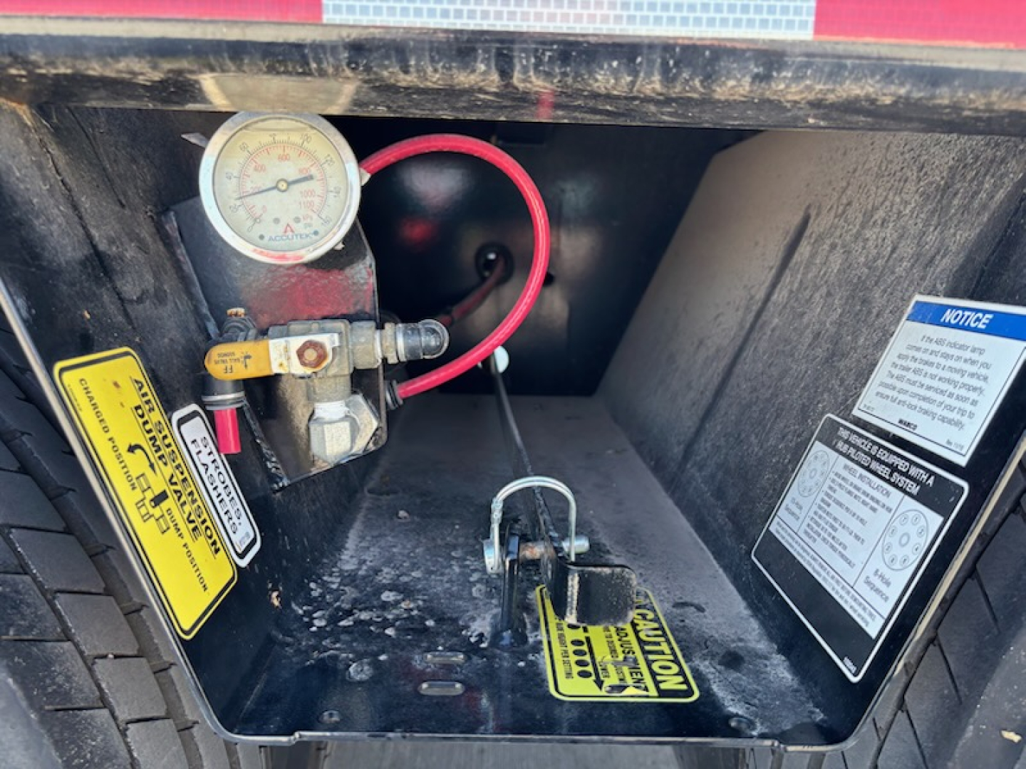 Trailer jack/air gauge panel showing a pressure gauge, red hose, control valve, and warning labels inside a toolbox area of a trailer.