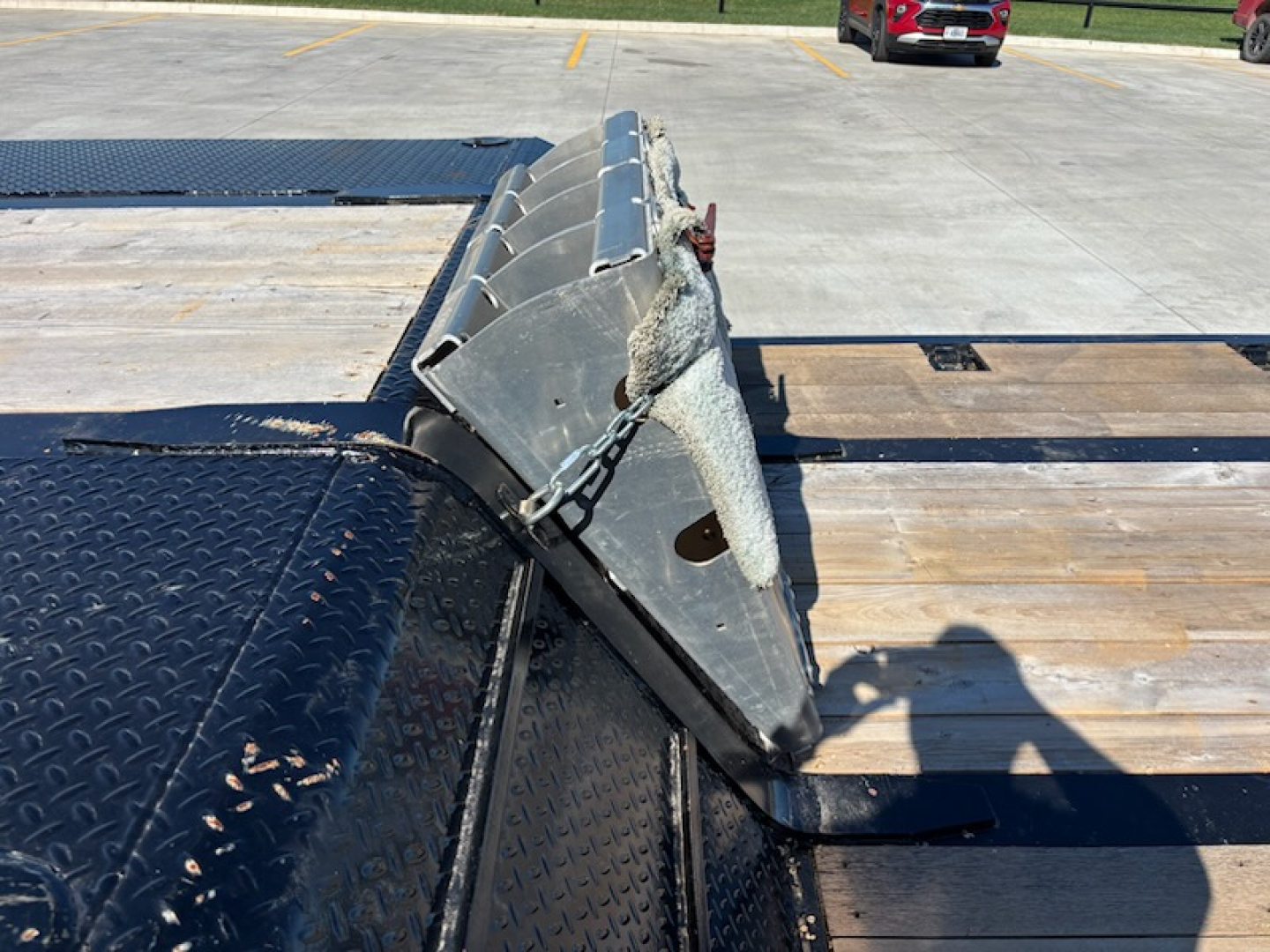 Trailer hitch and metal ramp/brace assembly on a flatbed trailer showing a chain and hinge mechanism with diamond plate surface