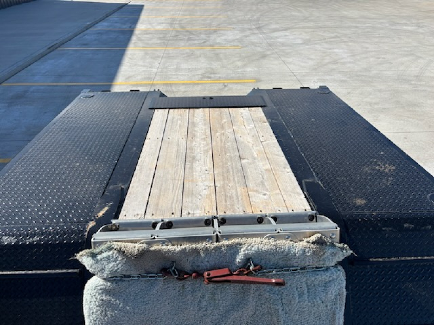 Rear view of a flatbed trailer with a wooden center deck and diamond plate sides. Visible are a small gray winch/strap device and a light-colored, textured guard near the back. The trailer bed has black diamond plate surfaces and a wood pla