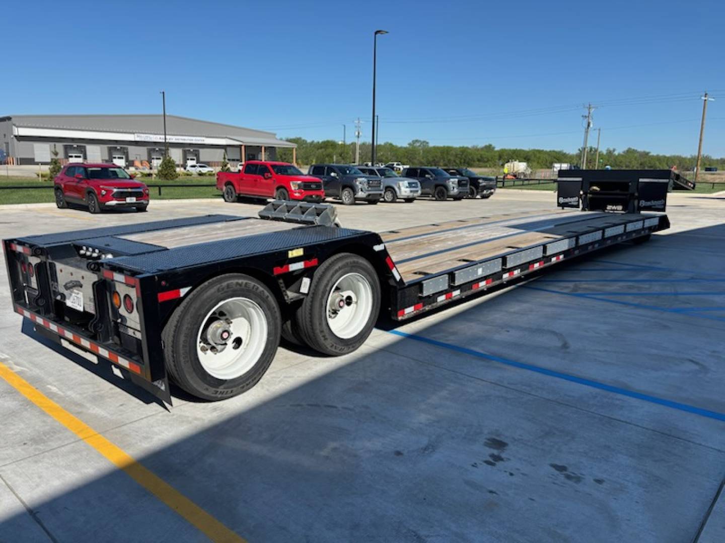 Flatbed trailer with dual rear axles and wooden deck. Visible text includes no obvious brand markings on the trailer; scene shows parking lot with several vehicles in background.