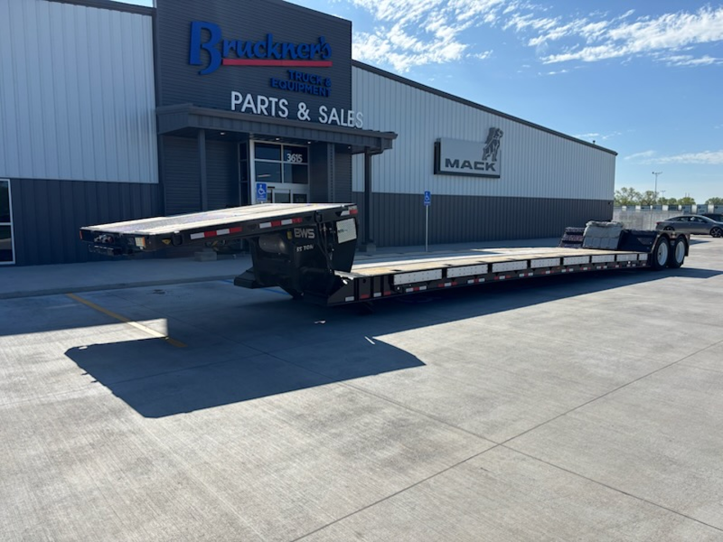 Flatbed trailer parked in front of a parts and sales building with Mack logo. Long low-profile trailer with metal decking and dual rear axles, visible trucks and trailers lot.