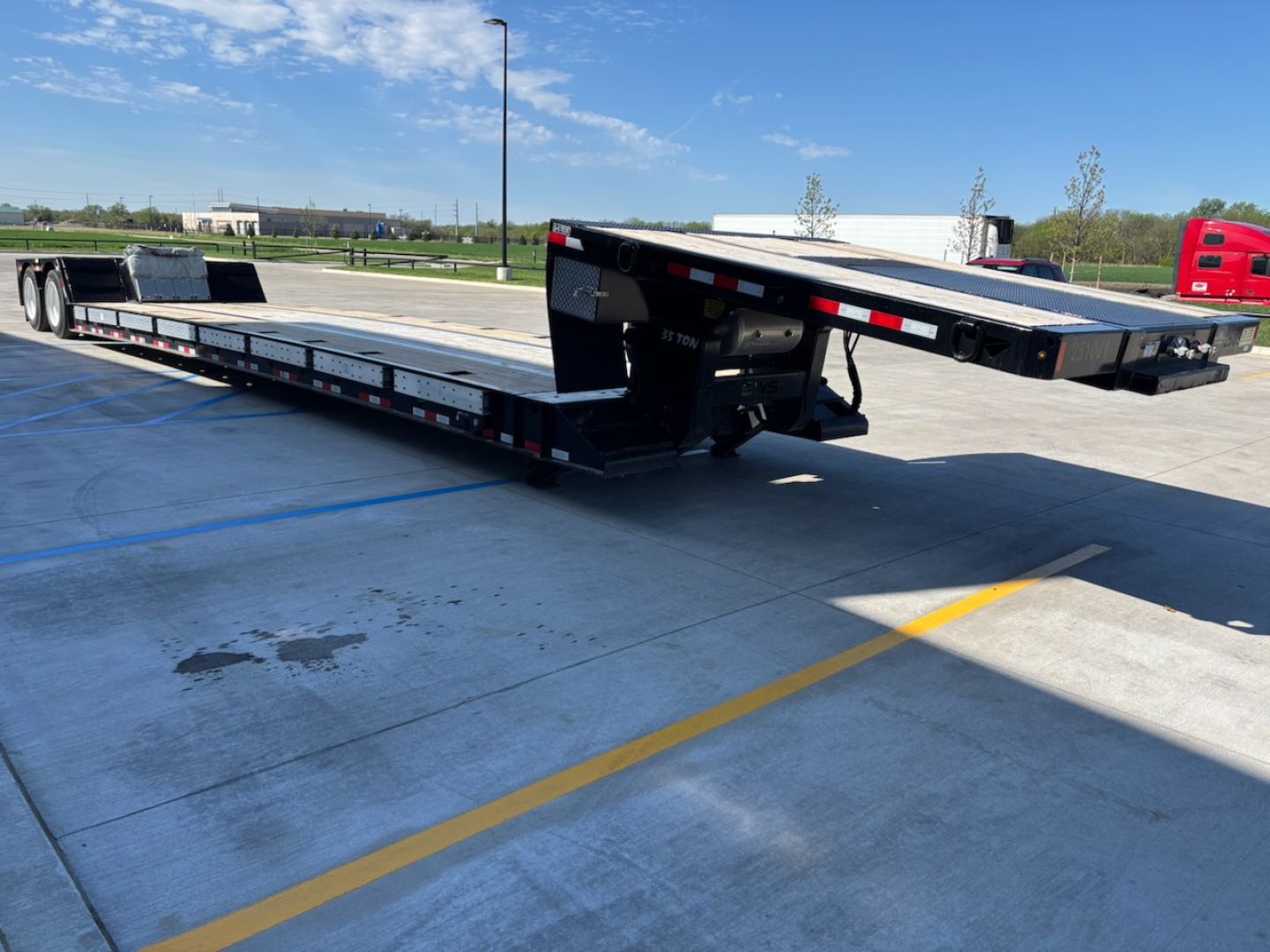 Flatbed trailer showing a low-profile gooseneck with loading ramps and reflective safety tape along the sides.