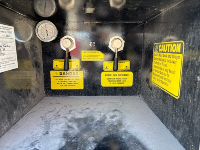 Trailer interior showing two deck lock levers with yellow warning labels, gauge, and caution stickers. Labels read: DANGER, DECK LOCK PIN WELVE? (text partially obscured). Caution sticker present on right with bold 'CAUTION'. Left label wit
