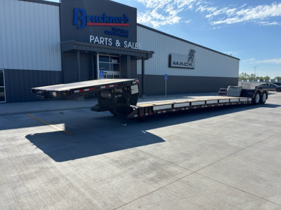 Flatbed trailer parked in front of a parts and sales building with Mack logo. Long low-profile trailer with metal decking and dual rear axles, visible trucks and trailers lot.
