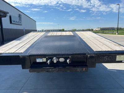 Flatbed trailer rear view showing platform with wooden deck and heavy-duty steel frame, hydraulic controls, and ramp ends.