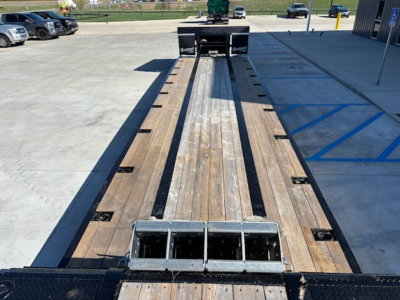 Flatbed trailer with wooden deck and metal ramps, viewed from rear, showing central steel channels and slotted tie-downs.