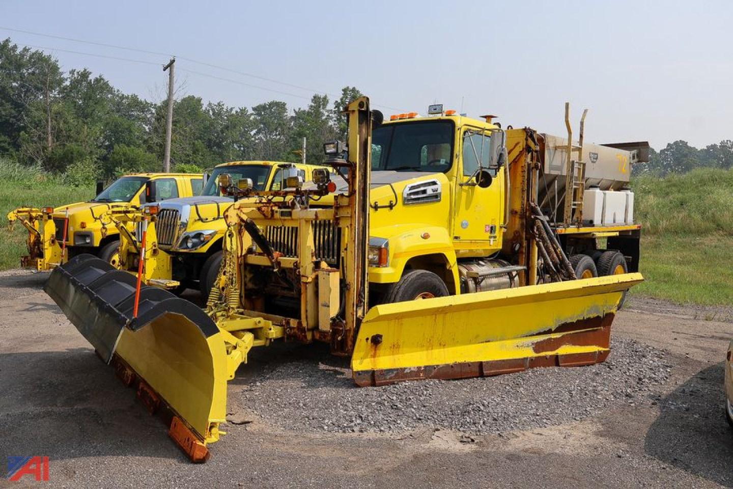 Western Star 4700 SF stake truck with plow and spreader in a lot; yellow maintenance truck with large front plow and side equipment.