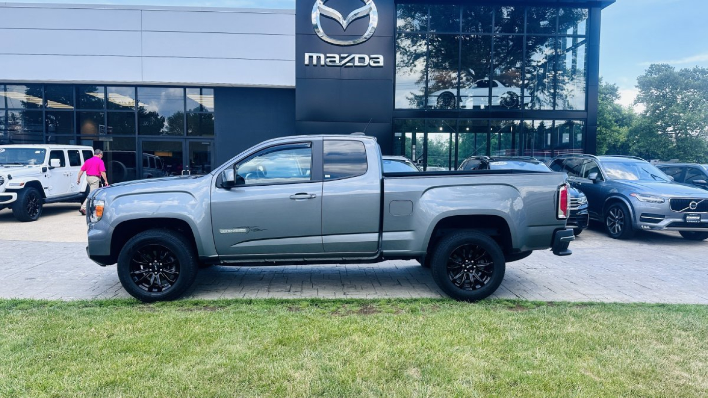 2021 GMC Canyon pickup truck in front of Mazda dealership showing side profile with crew cab, gray exterior, black wheels, and stock number plate visible.