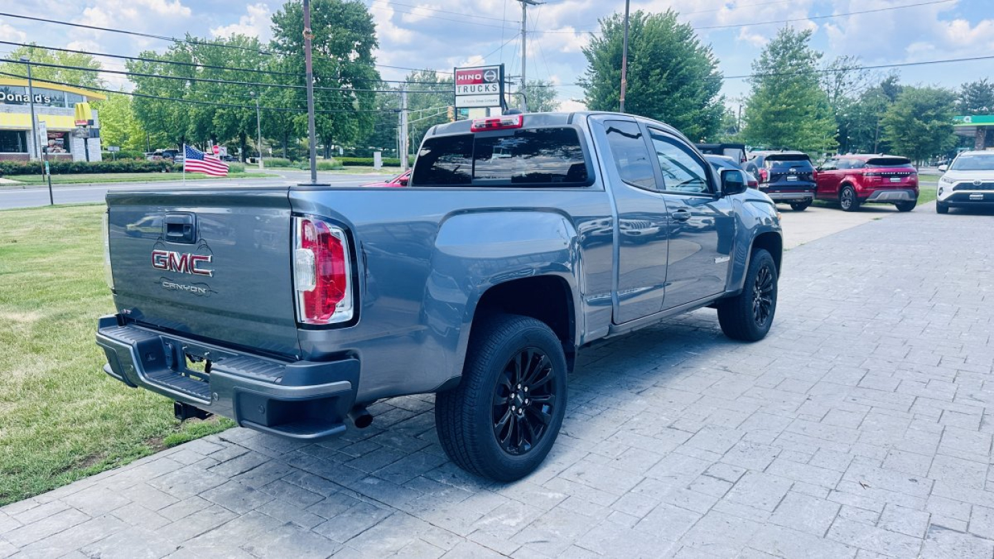 2021 GMC Canyon pickup truck rear-right view showing tailgate with GMC badge, 'CANYON' label, black wheels, light blue-gray color, dealership lot with other vehicles in background.