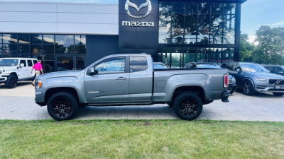 2021 GMC Canyon pickup truck in front of Mazda dealership showing side profile with crew cab, gray exterior, black wheels, and stock number plate visible.