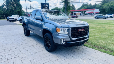GMC Canyon pickup truck in a light blue color, front three-quarter view, parked on a paved lot near a gas station.