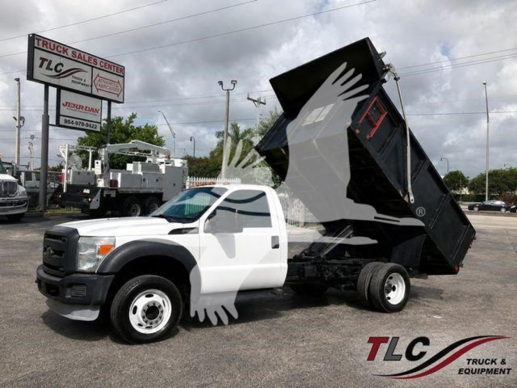 Truck bed raised in dumping position on white 2014 Ford F-450 XL dump truck, side view, TLC Truck & Equipment lot in background.