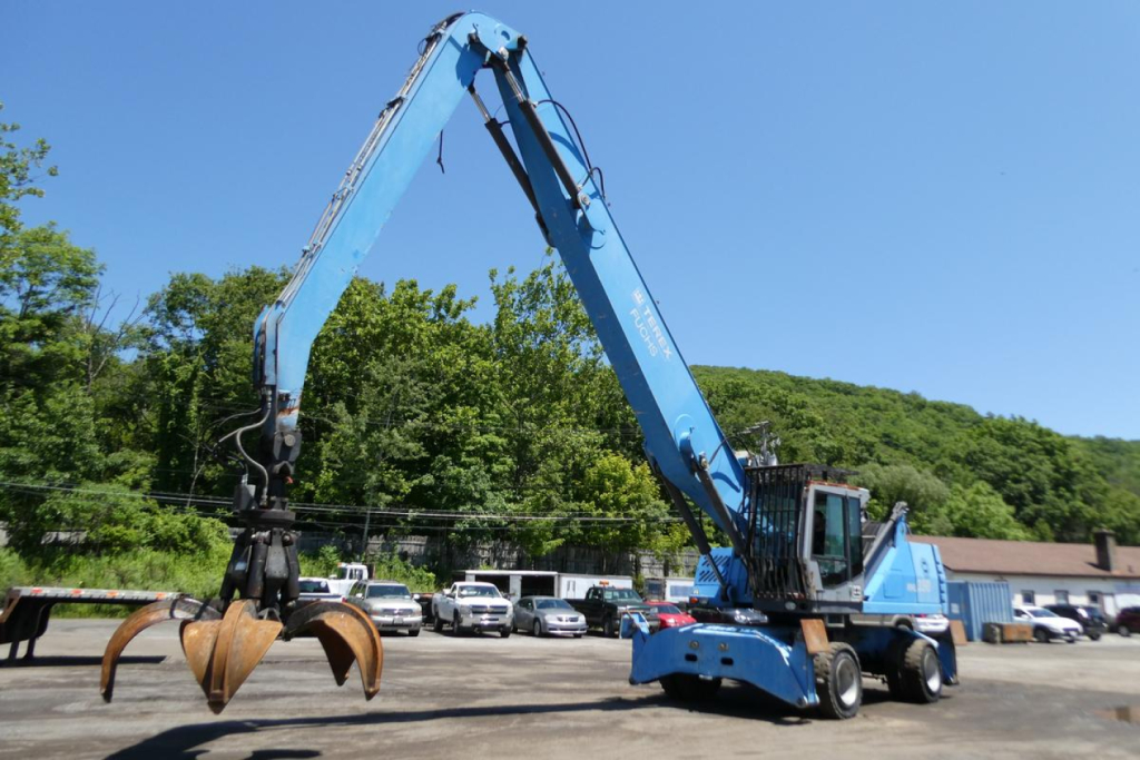 Terex MHL350D material handler in operation view showing long blue boom with clamshell grapple at end; operator cab and outriggers; background trees and parked vehicles.