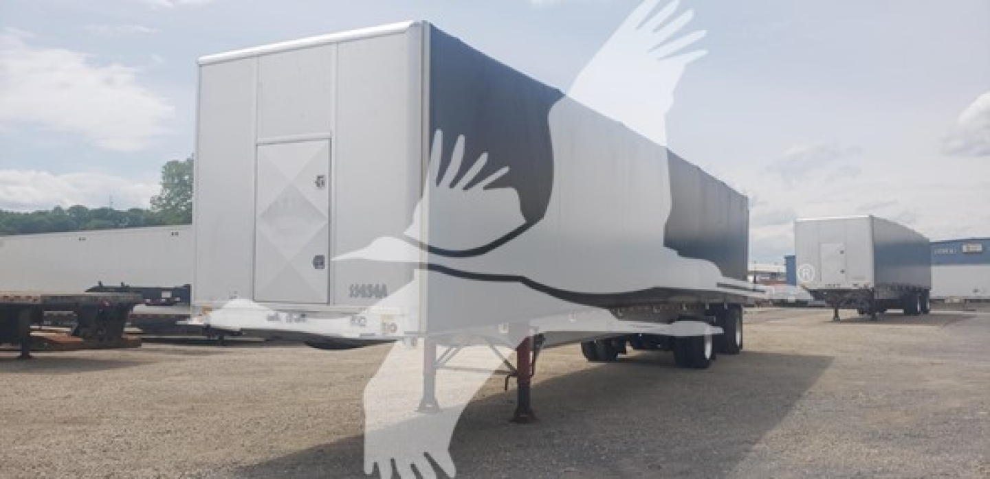 Trailer in a yard showing a curtainside semi-trailer with a closed rear door and side panels, under clear sky.