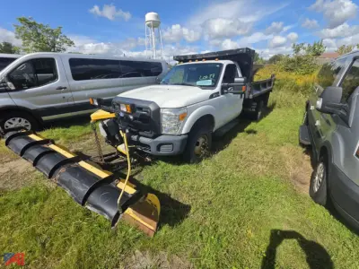 Ford F-350 Super Duty dual rear wheel truck with plow blade attachment in grassy lot; white cab with black snow plow mount, chrome grille, and auxiliary lights.