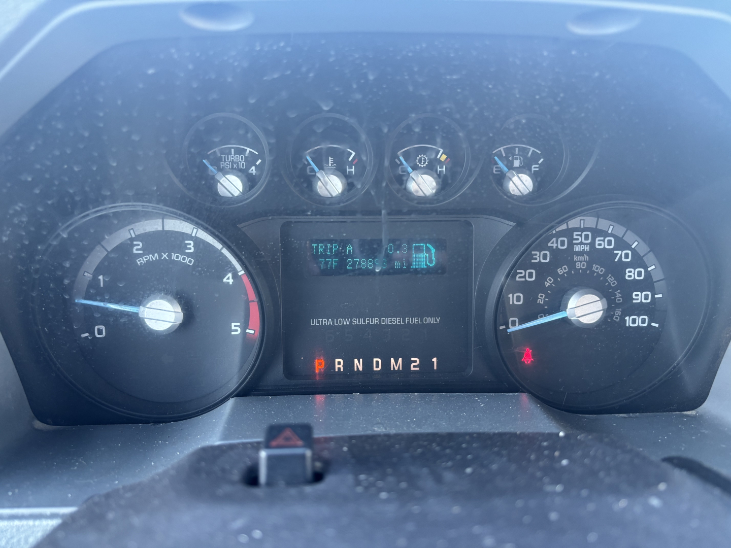 Instrument cluster of a 2012 Ford F-550 dash showing multiple gauges and digital display with transmission gear indicator PRNDM21 and fuel/engine readings.