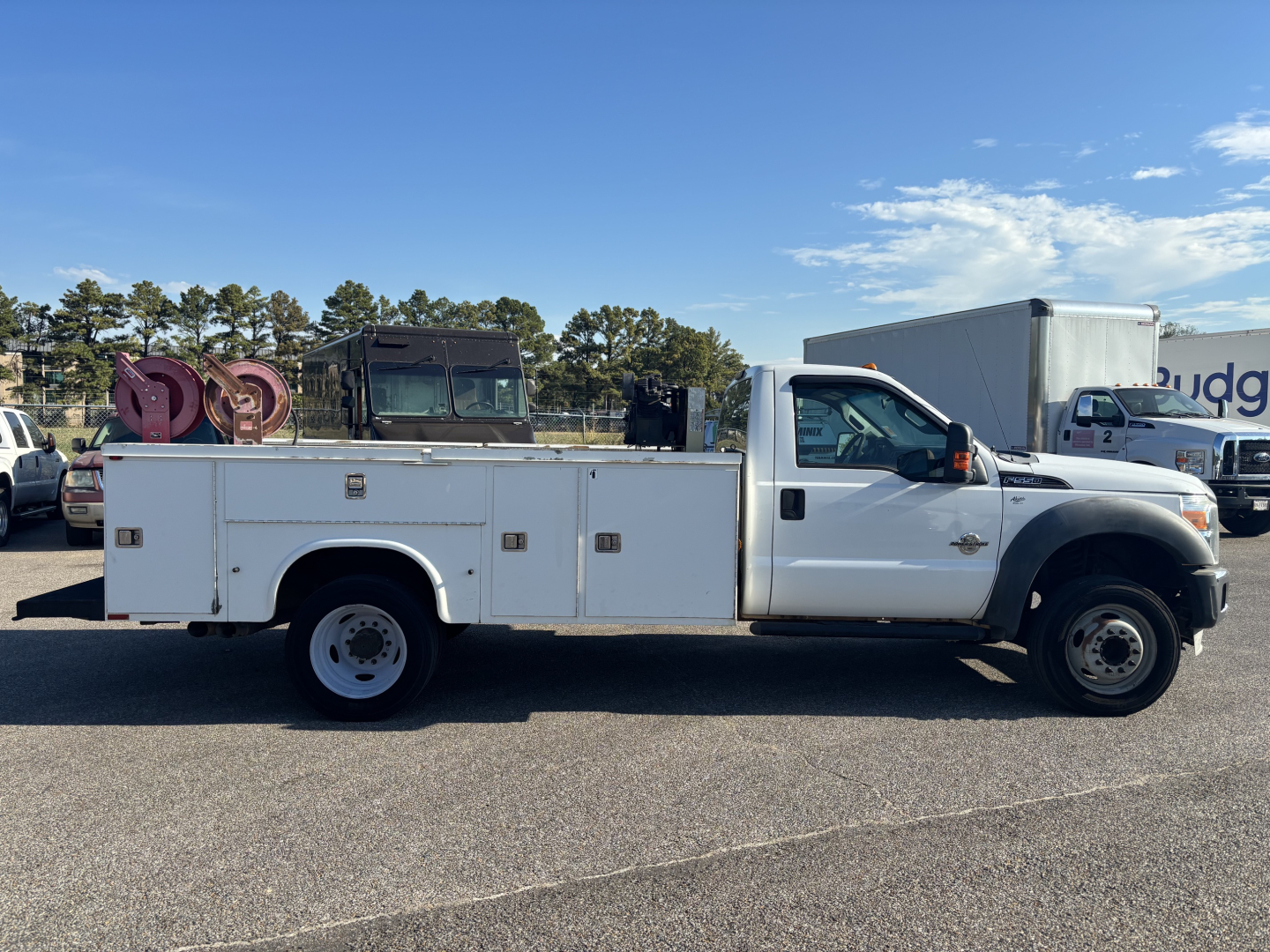 Ford F550 utility/service bucket truck side view showing utility bed with multiple compartments and a rack at rear. White cab and chassis with long wheelbase in a parking lot.