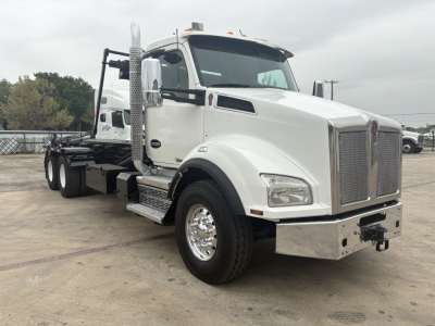 Kenworth T880 roll-off truck in white, front-right view showing cab, chrome grille, tall exhaust stack, side steps, and rear chassis for wheel-lift bed.