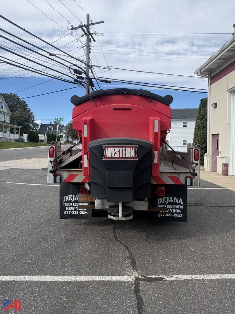 Rear view of a red dump truck hopper with Western plow/sander equipment. Visible text includes 'WESTERN' on the hopper and contact numbers on the mud flaps: 'DEJANA TRUCK EQUIPMENT' and phone numbers. Utility poles and a street in backgroun