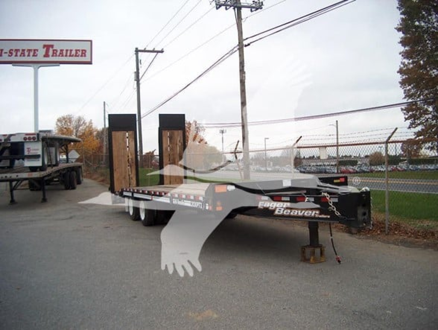 Flatbed trailer with dual ramps and Eager Beaver branding, parked outdoors near a chain-link fence and sign, TL088527 stock number.