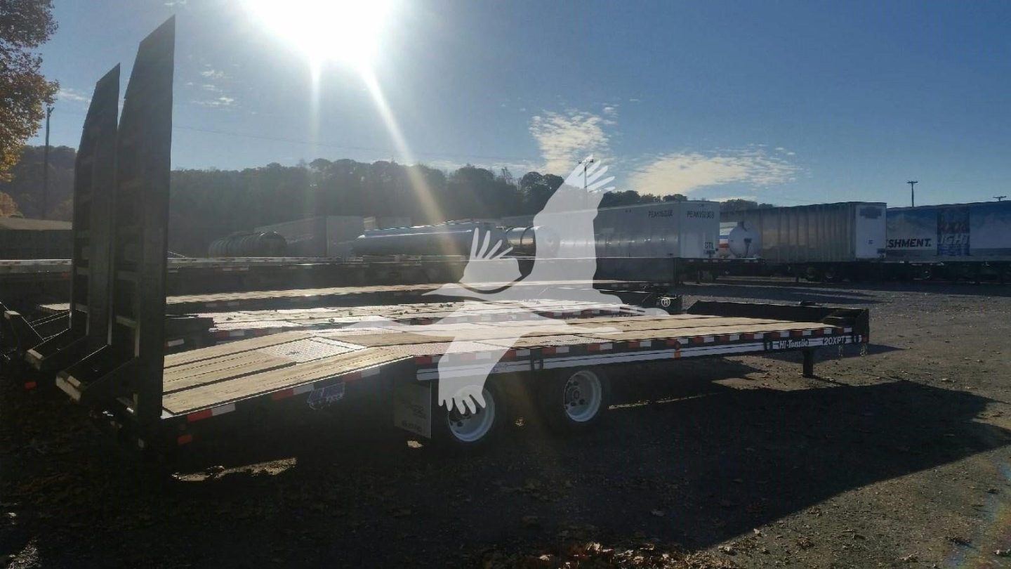 Flatbed trailer in a yard with dual axles and ramp up, showing stamped metal deck and side rails, under clear sky.