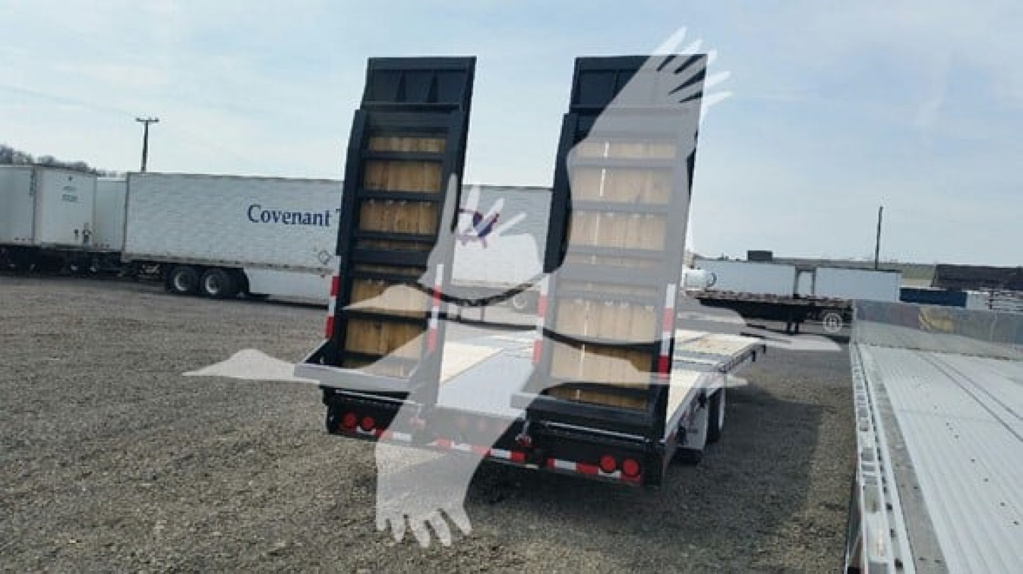 Flatbed trailer with post ramps in open lot, dual rear ramps raised, wooden deck visible, black frame, tail lights at back, other trailers in background.