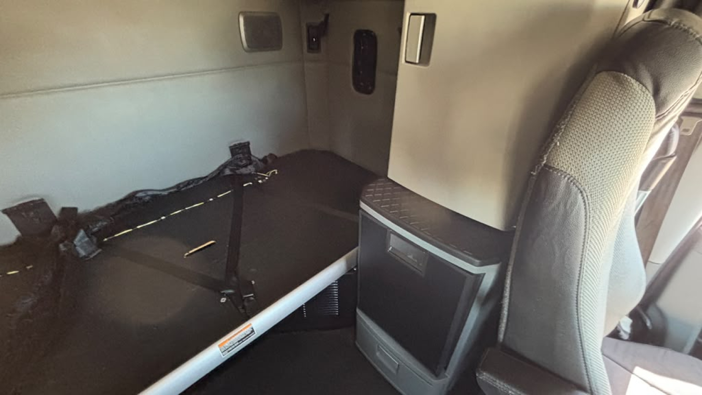 Kenworth T680 cab interior showing sleeper area with cushions and small TV or monitor mounted on cabinet; seat and side panel visible.