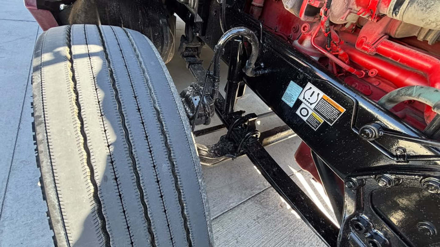 Kenworth truck chassis and front axle showing large dual tires and undercarriage with black metal frame and red engine components in background.