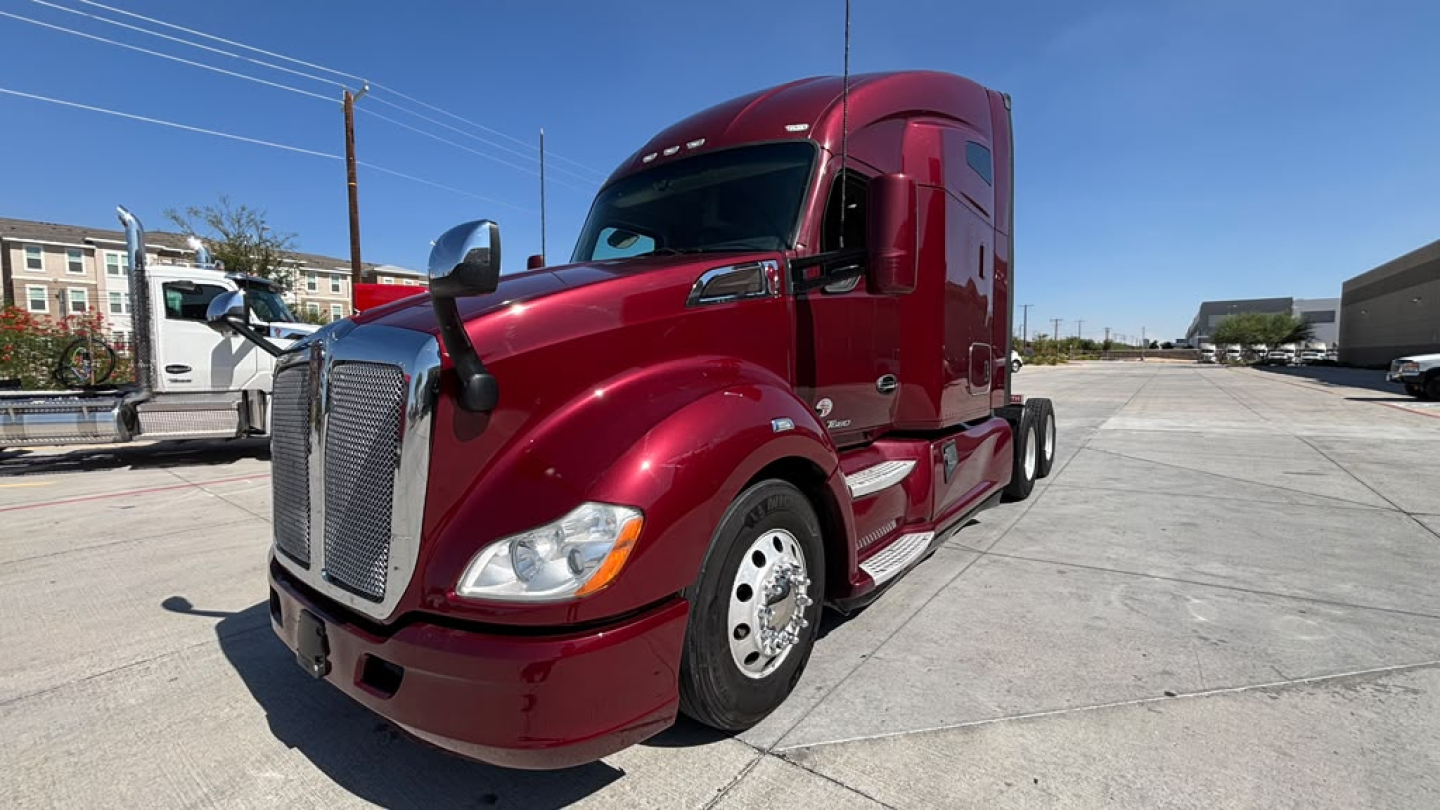 Kenworth T680 with deep red exterior, front left side view showing sleeper cab, chrome front grille, headlight, and large fuel/air tanks; highway in background.